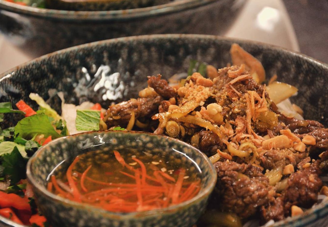 A close-up of wok-tossed beef with lemongrass, crushed peanuts and crispy fried shallots over fresh salad, served with a bowl of nước chấm dipping sauce and shredded carrot.