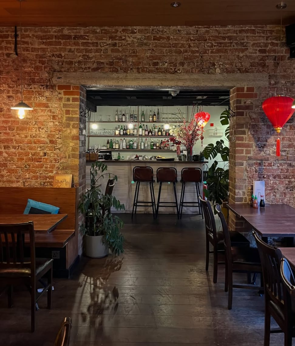 The Việt Garden dining room — exposed brick walls, dark wooden tables and chairs, a pendant light, red silk lanterns and a stocked bar visible through an archway at the back.
