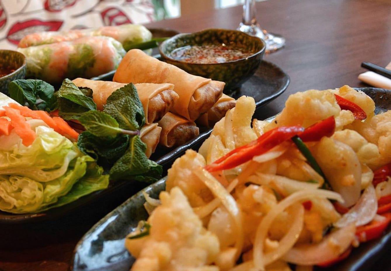 A sharing spread on dark ceramic plates — fresh summer rolls with prawns, golden crispy spring rolls with mint, salt and pepper squid with chilli and onion, and a bowl of dipping sauce.