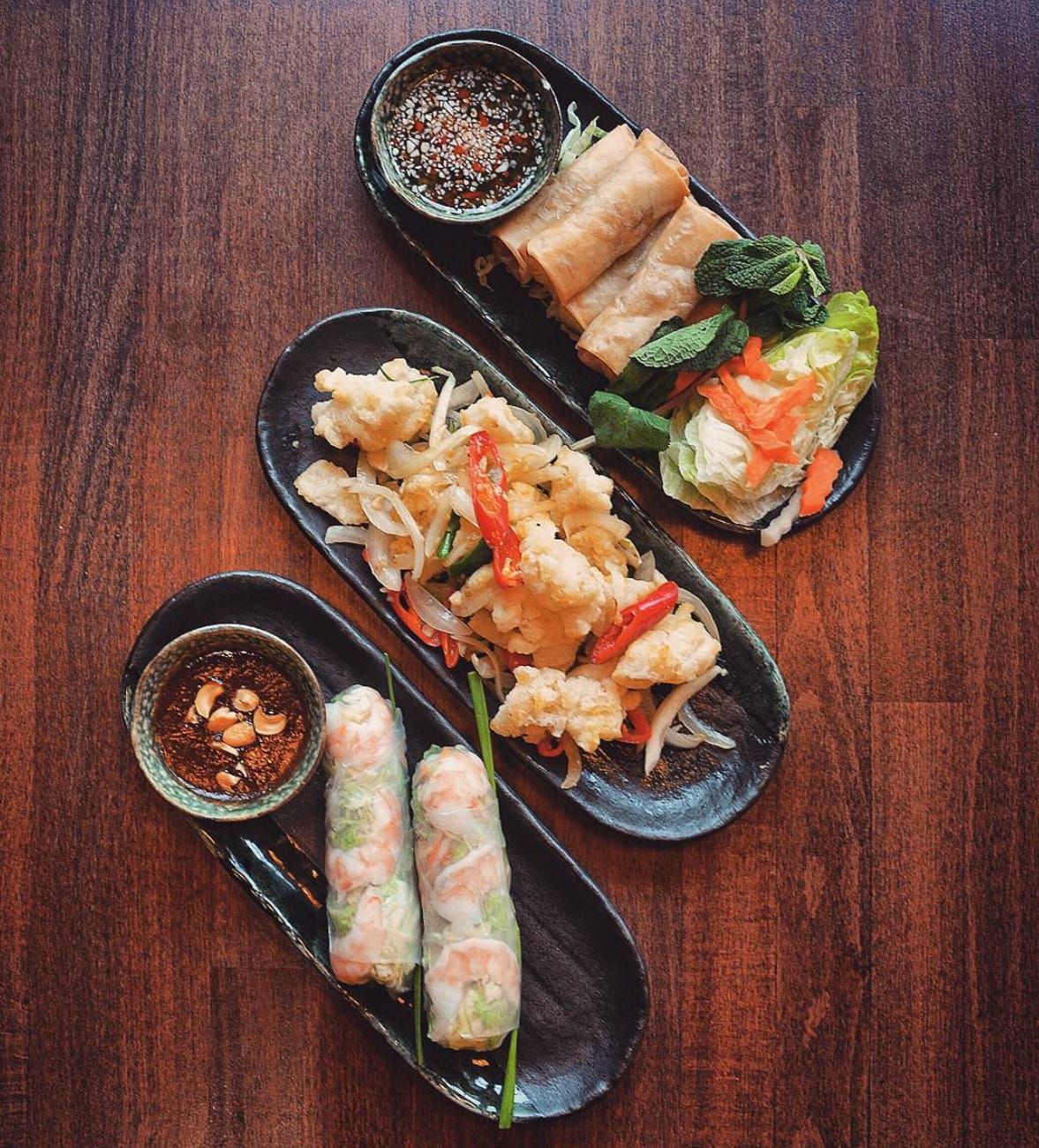 Overhead view of three long dark plates on a wooden table — crispy spring rolls with herbs and pickled carrot, salt and pepper squid, and fresh prawn summer rolls with a peanut dipping sauce.
